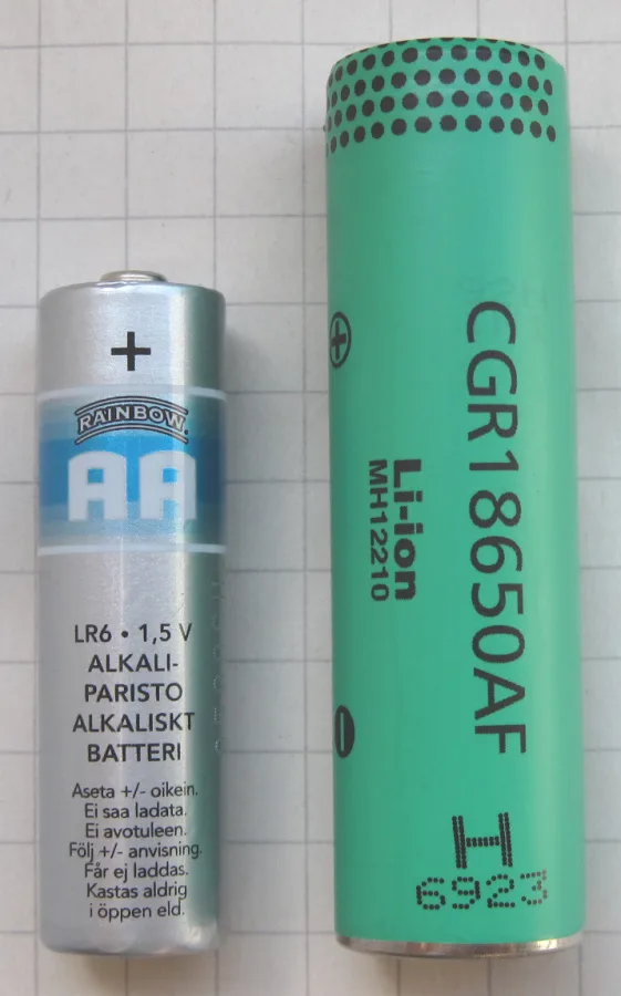 Kareli kâğıt üzerinde yan yana duran gümüş renkli bir AA alkalin pil ve ondan daha kalın ve uzun, yeşil bir 18650 lityum iyon hücre