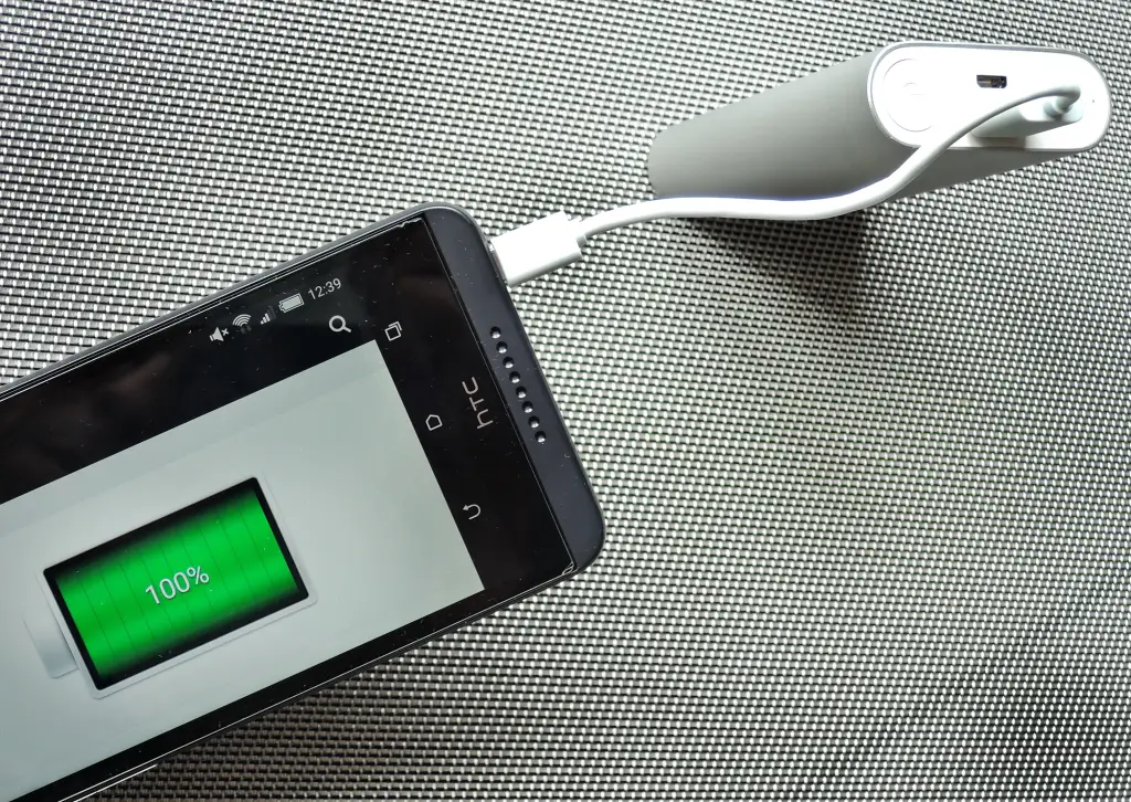 Gri dokulu bir zemin üzerinde beyaz kabloyla silindir biçimli beyaz bir powerbank'a bağlanmış, ekranında yüzde 100 şarj gösteren akıllı telefon